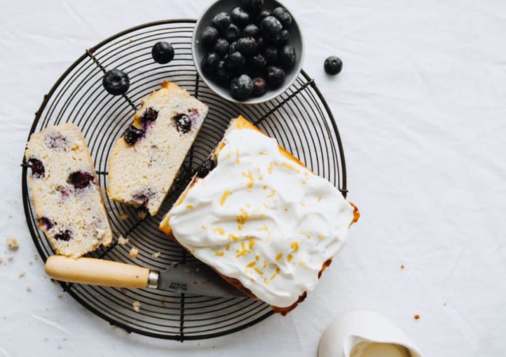 A loaf of blueberry cake with icing, sliced, on a metal cooling rack with a bowl of blueberries and a jug of milk nearby, on a white cloth.