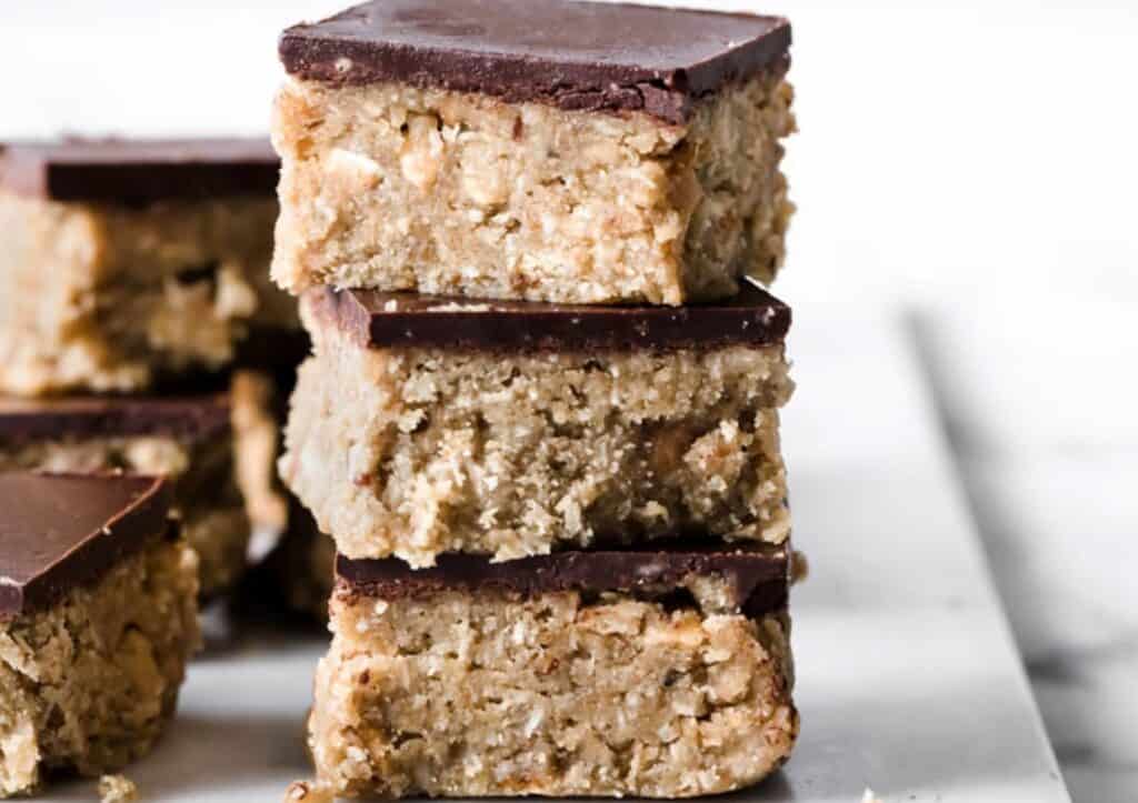 Stacked homemade scotcheroos with a thick chocolate layer on top, displayed on a white surface.