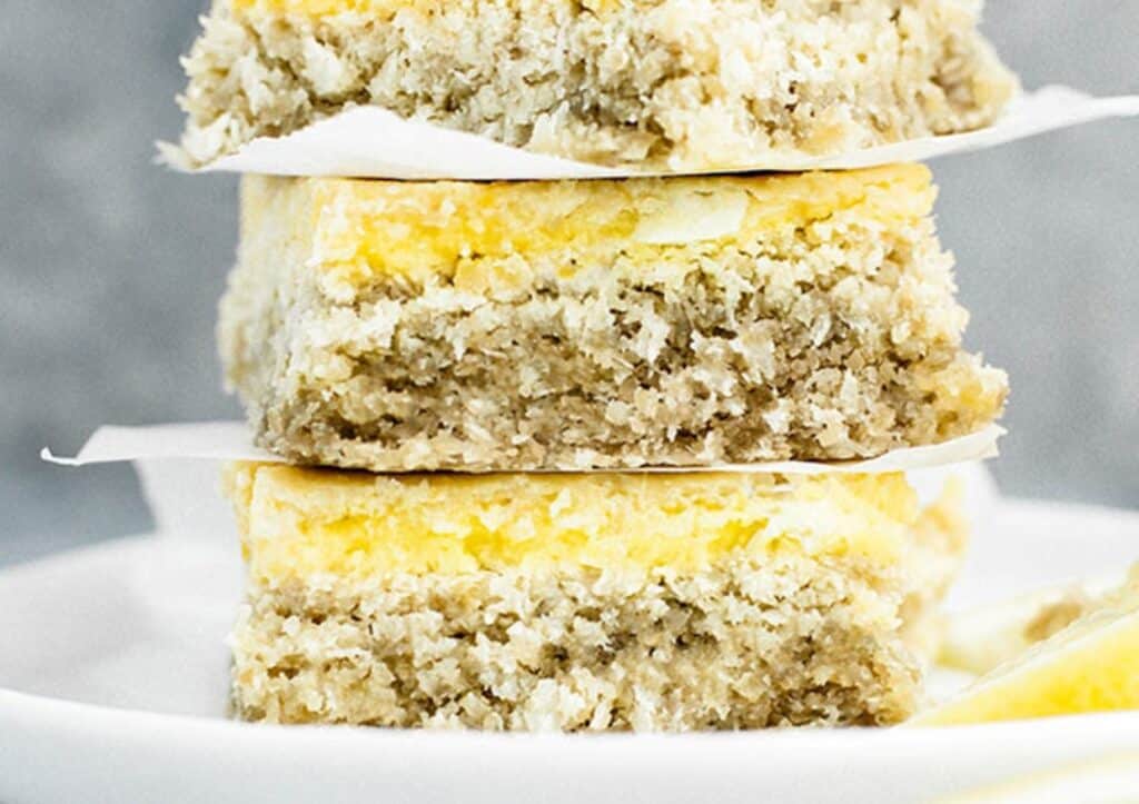 A close-up image of a stack of coconut-crusted lemon bars on a white plate, showing distinct layers and a textured topping.