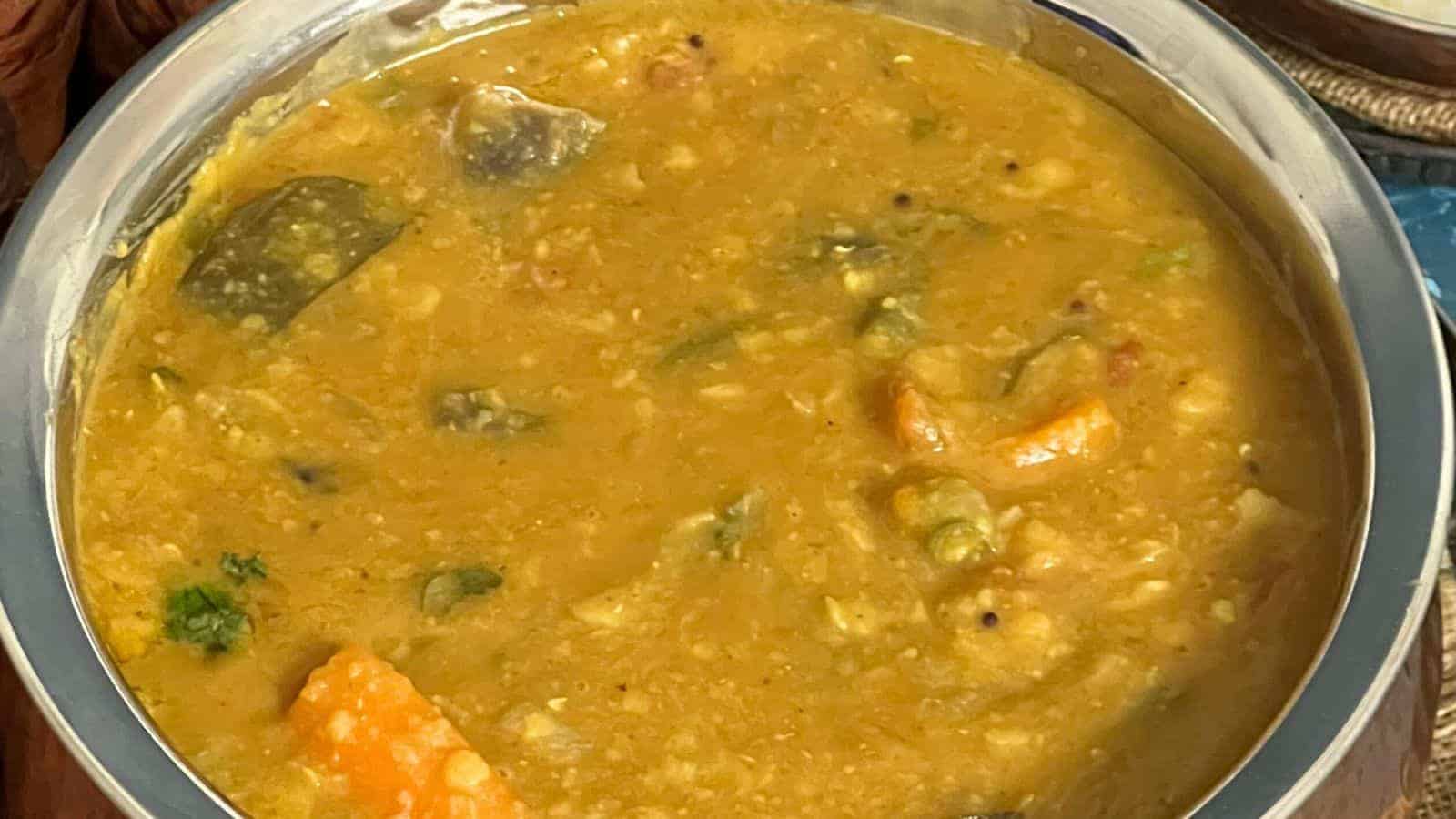 A bowl of sambar, a traditional south indian lentil soup with mixed vegetables and herbs, served in a stainless steel bowl.