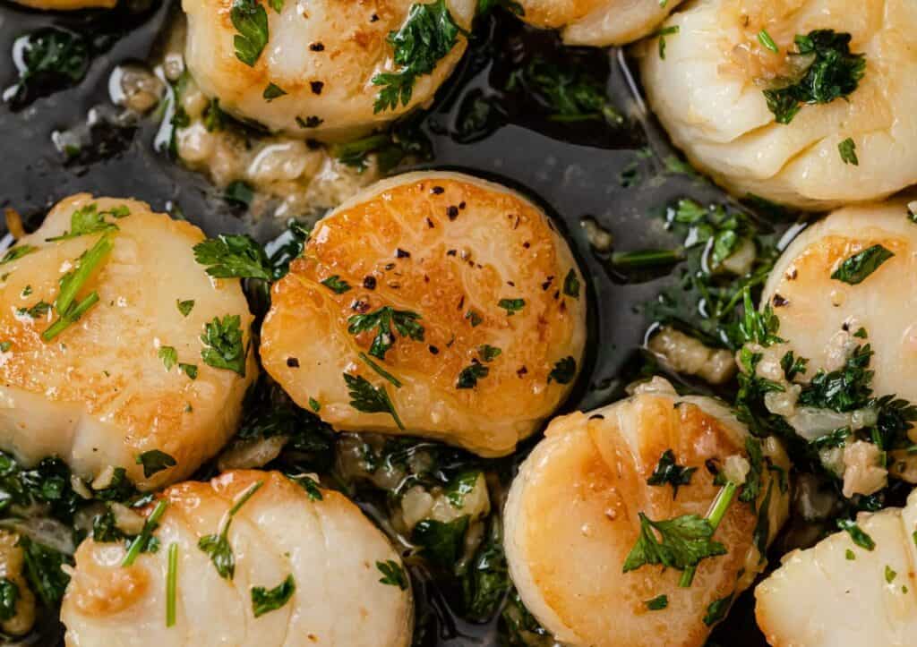 Seared scallops garnished with chopped parsley in a black skillet.