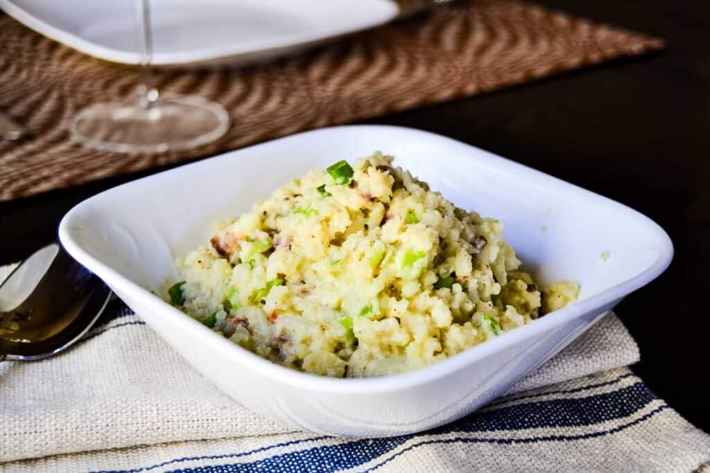 A bowl of mashed cauliflower with chives on a wooden table with a spoon and a wine glass in the background.