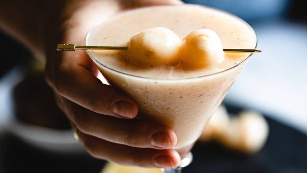 A close-up of a hand holding a frosted glass containing a creamy cocktail garnished with two lychee fruits.