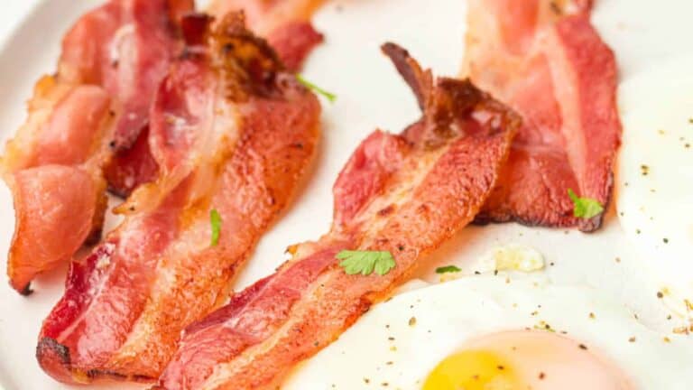 Close-up of crispy bacon strips and fried eggs on a white plate, sprinkled with herbs.
