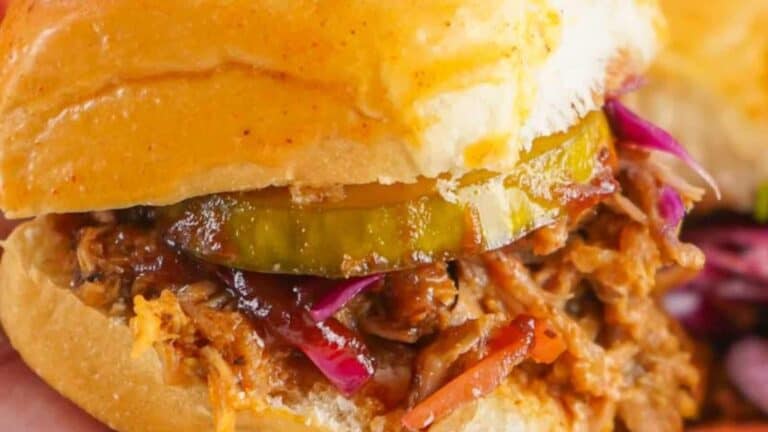 Close-up of a pulled pork sandwich with red cabbage, carrots, and pickles on a bun, showcasing its juicy filling and fresh toppings.