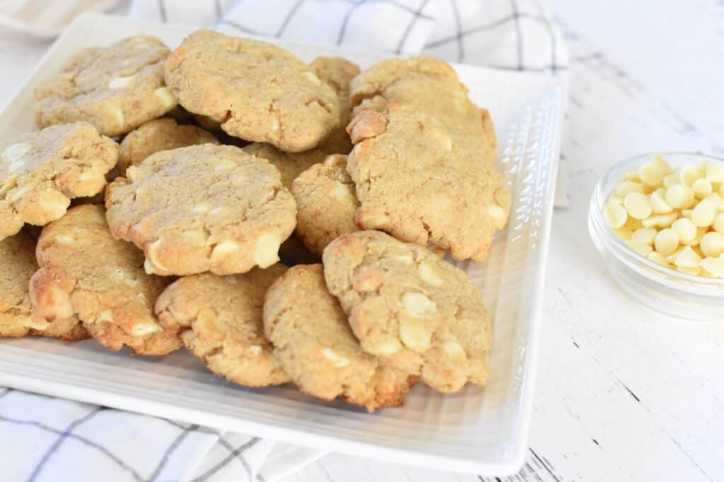 A plate of homemade cookies with white chocolate chips next to a small bowl of additional white chocolate chips.