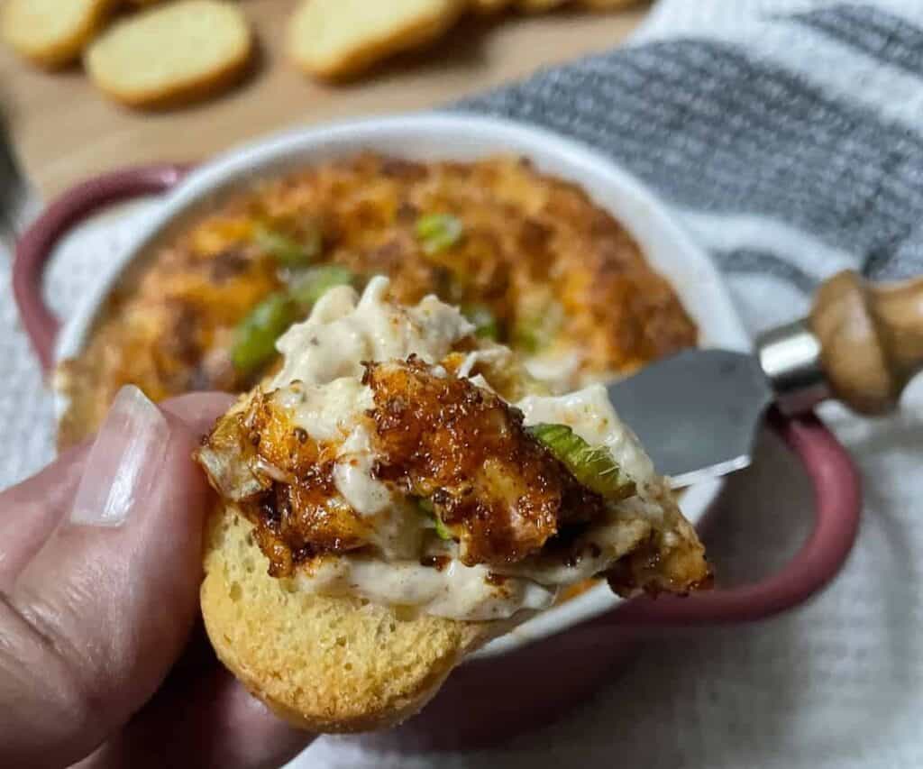 A close-up image of a hand holding a cracker topped with a scoop of creamy crab dip and chopped green onions, with a bowl of the dip in the background.