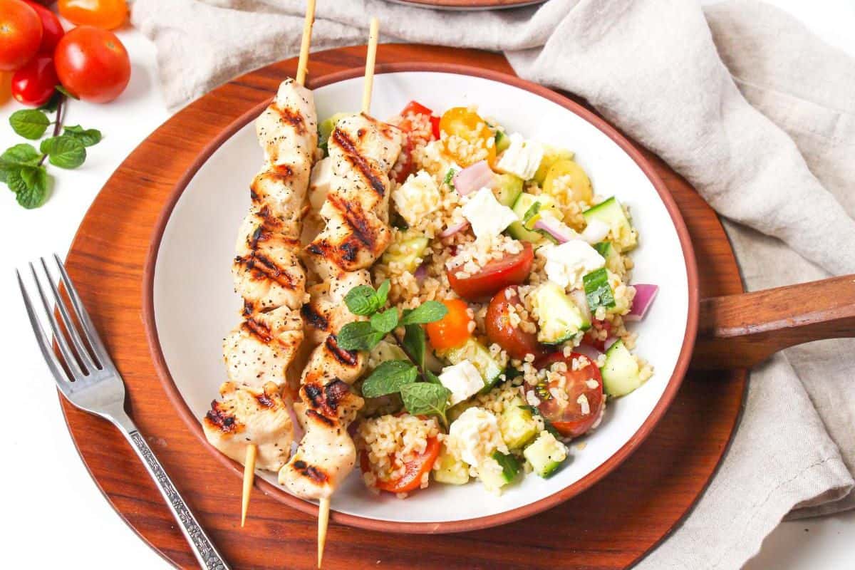 A white plate with tomato and cucumber bulgur salad topped with feta cheese next to two grilled chicken skewers.