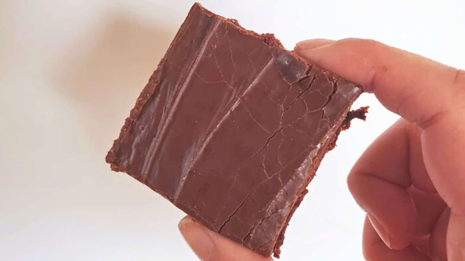 Image shows A hand holding a chocolate covered square brownie with a cracked surface against a light background.