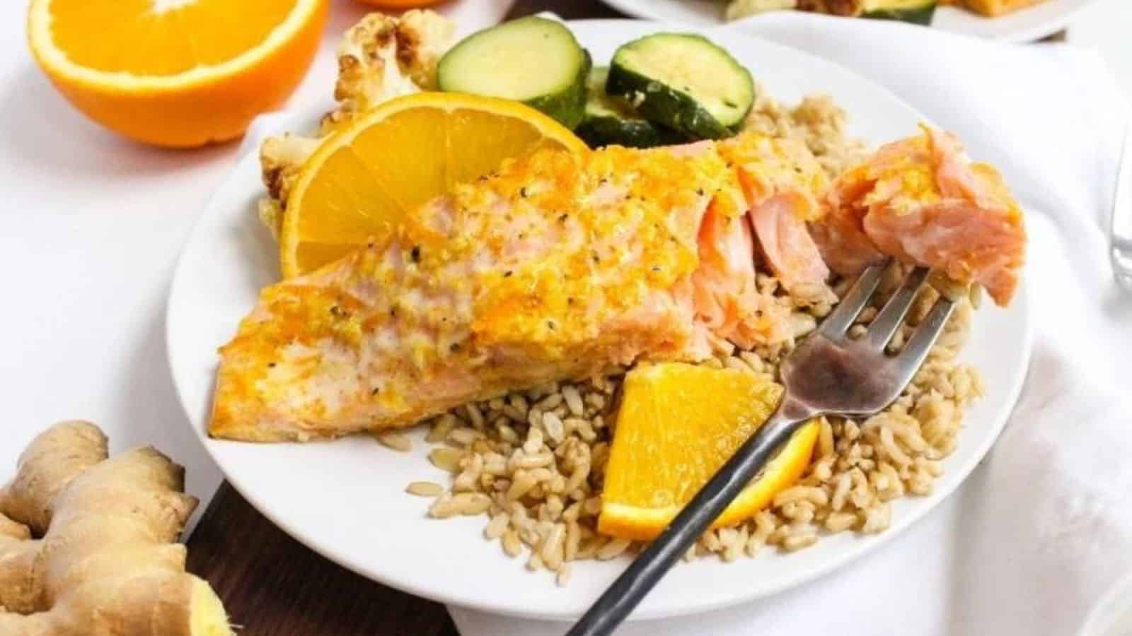 Flaky salmon topped with orange ginger glaze on a white plate with brown rice, roasted zucchini slices, and orange slices next to a fork.