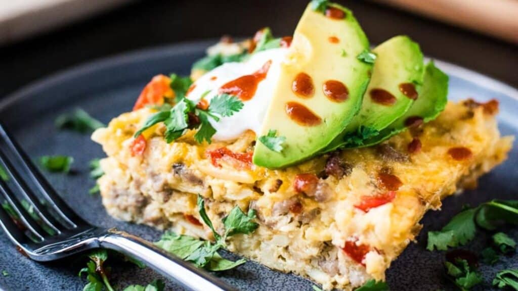 A slice of layered mexican casserole topped with avocado slices and sour cream, garnished with fresh cilantro, served on a black plate.