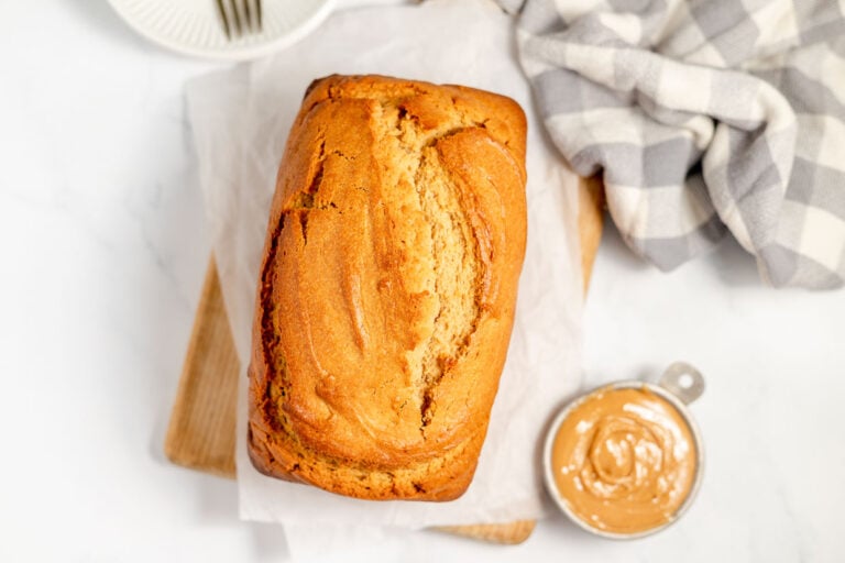 Top view of a freshly baked loaf of bread on a white surface with peanut butter in a small bowl and a striped cloth on the side.