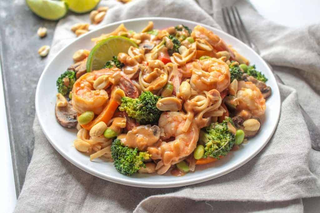 Shrimp, broccoli, bell peppers, mushrooms, and brown rice noodles drizzled with peanut sauce on a white plate.