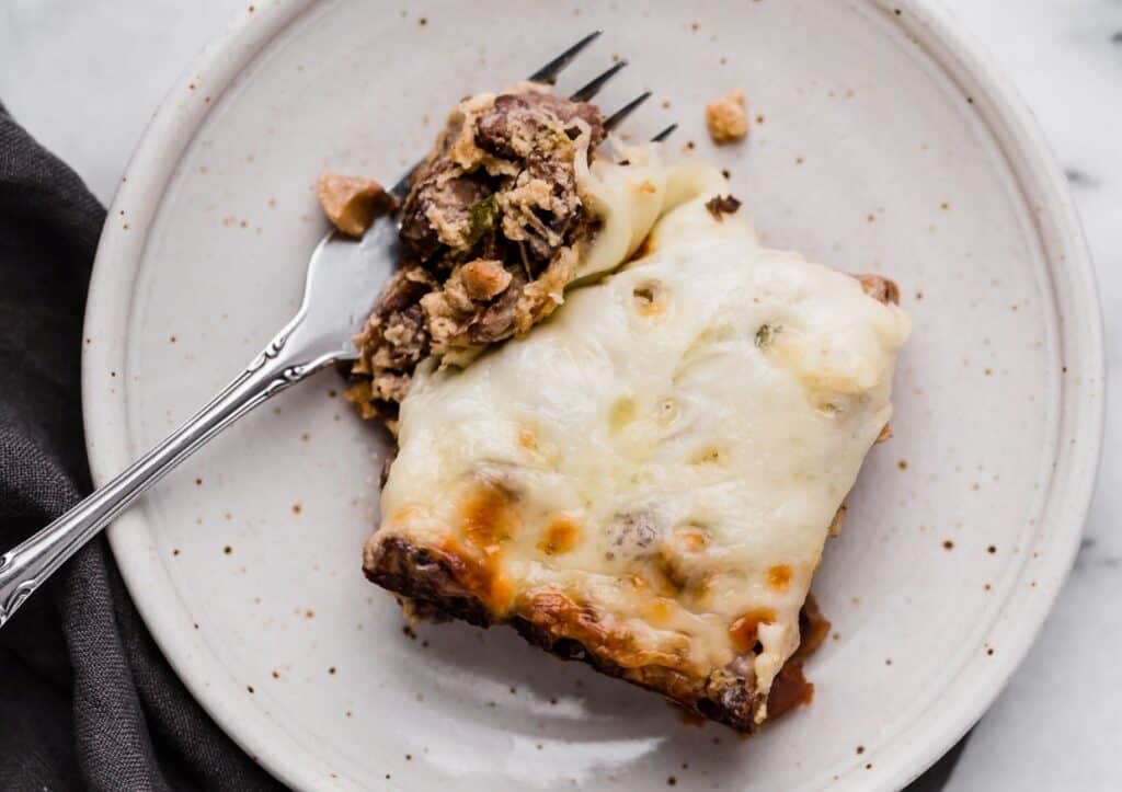 A slice of Philly cheese steak casserole with melted cheese on a plate, partially eaten with a fork resting on it.