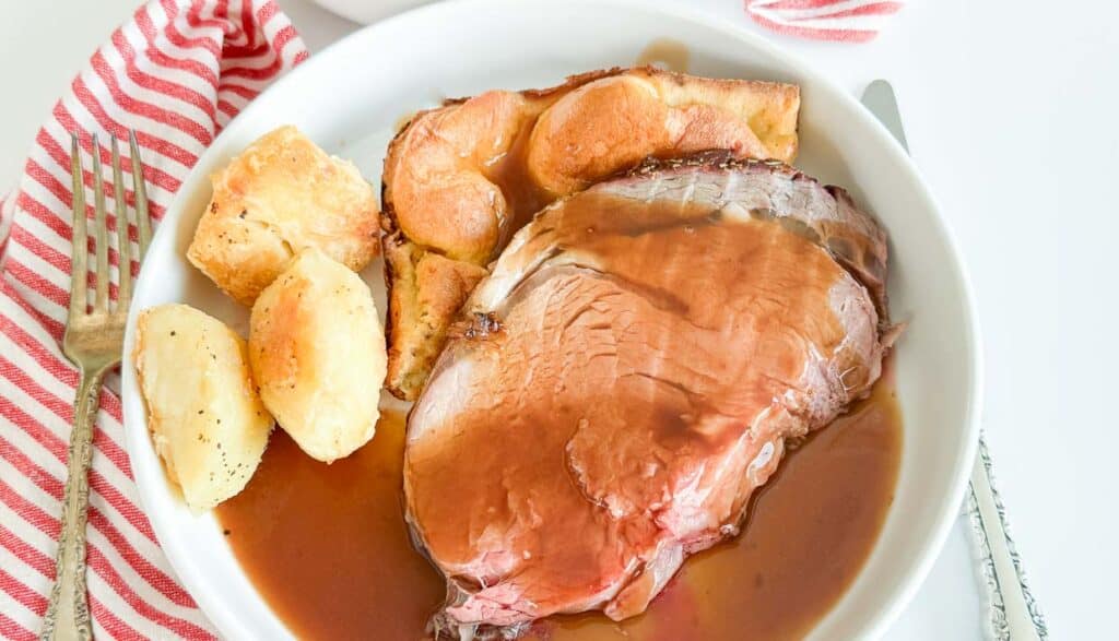 Sliced roast beef with gravy and roasted potatoes on a white plate, accompanied by a red and white striped napkin on the side.
