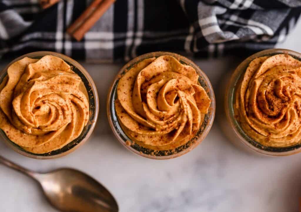 Three glass jars filled with creamy pumpkin mousse topped with a cinnamon swirl, accompanied by vintage spoons and a checkered napkin, on a marble surface.