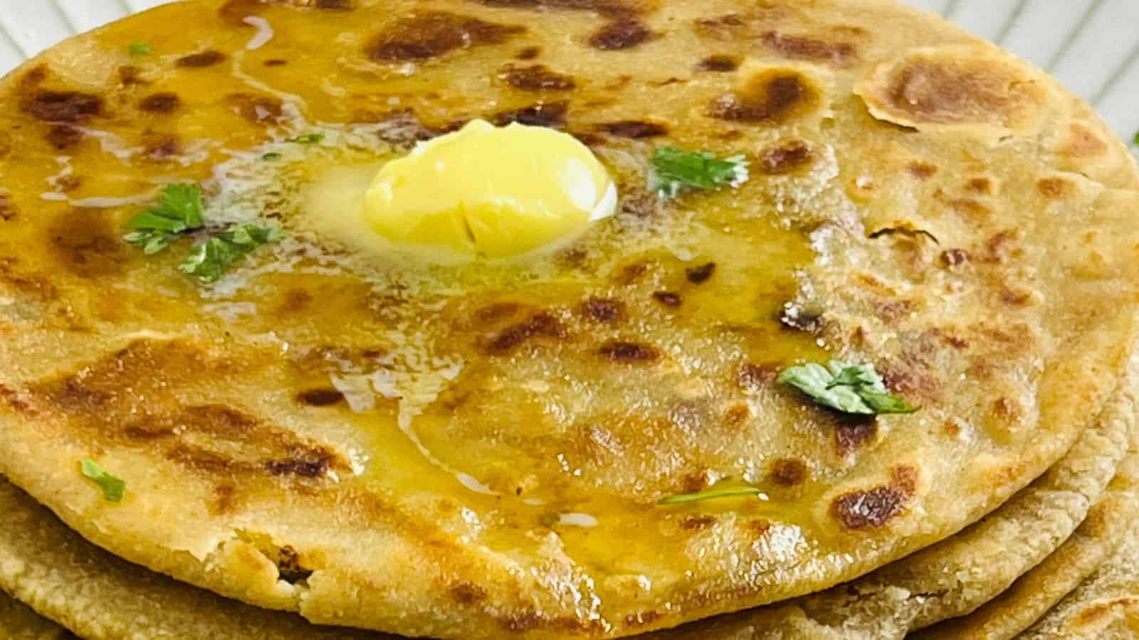Close-up image of aloo paratha, an indian stuffed flatbread, topped with a pat of melting butter and sprinkled with fresh herbs.