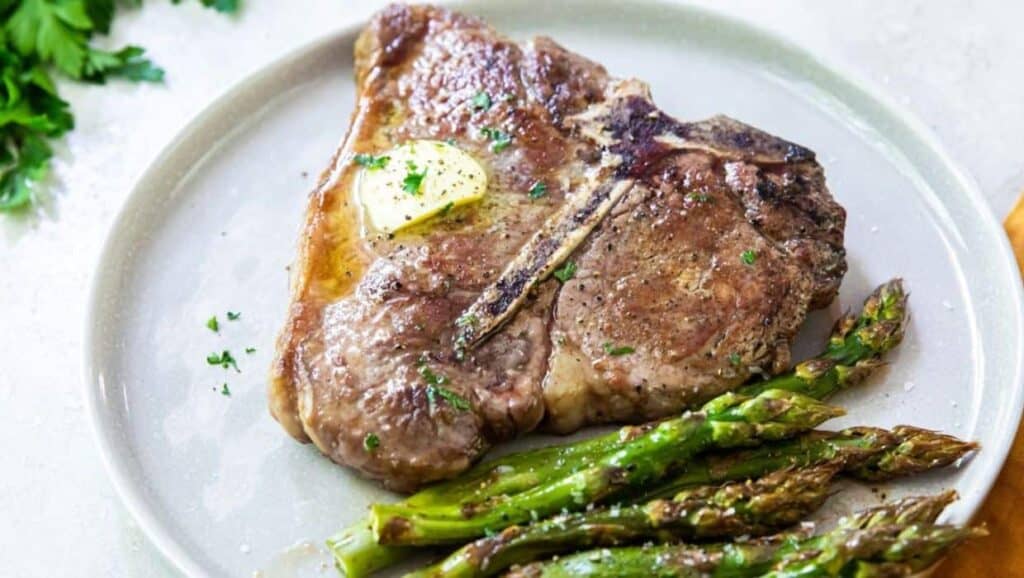 A grilled steak topped with butter and garnished with parsley, served with roasted asparagus on a white plate.