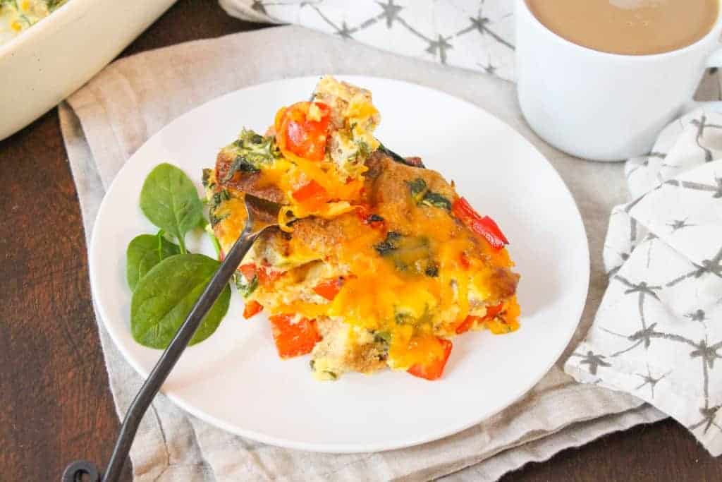 A slice of egg casserole with sausage, cheese, vegetables, and whole wheat bread on a white plate next to a mug of coffee.