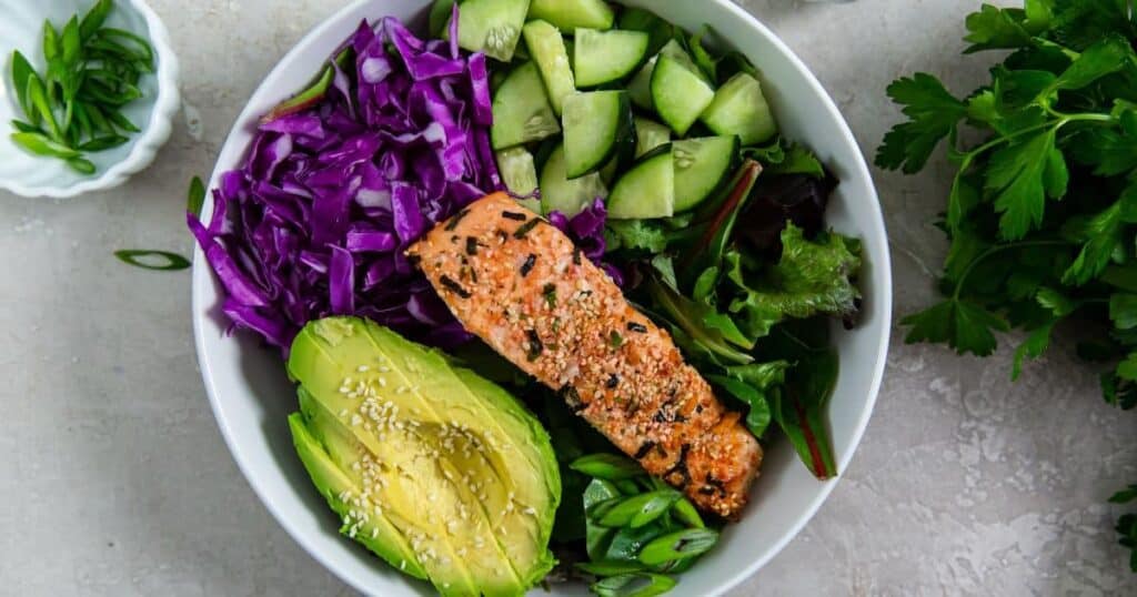 A healthy bowl featuring grilled salmon, sliced avocado, cucumber, red cabbage, and mixed greens, garnished with sesame seeds.