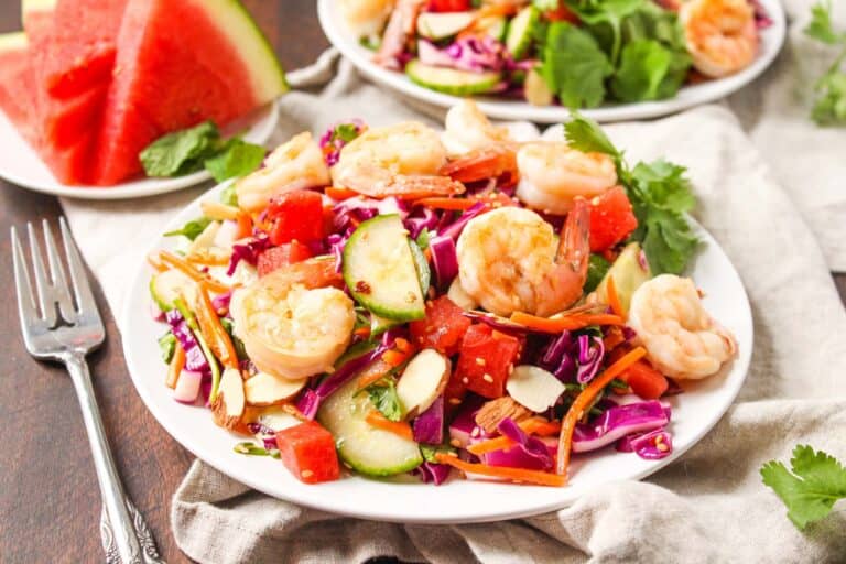 A colorful shrimp and watermelon salad with cucumbers, carrots, and leafy greens on a white plate, accompanied by a slice of watermelon.