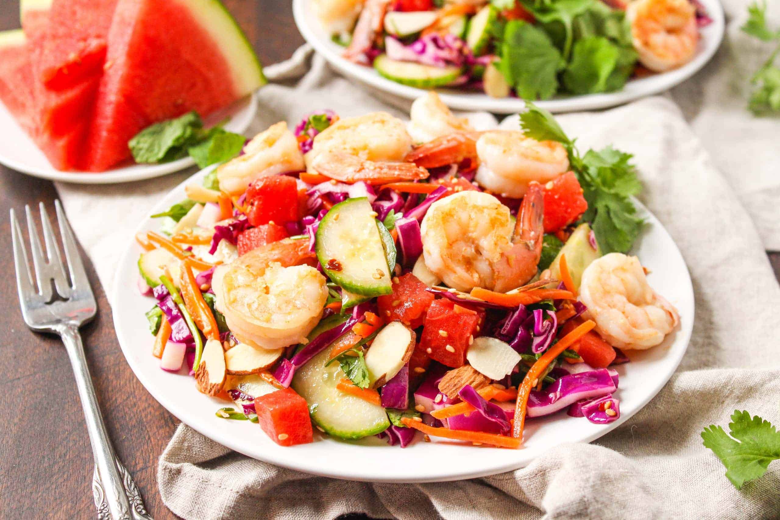 A white plate with shrimp, watermelon, cucumber, shredded cabbage, and grated carrots.