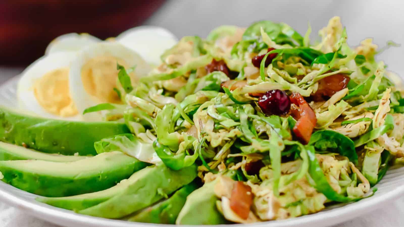 A serving of shaved brussels sprouts salad next to the serving bowl of shaved brussels sprouts salad with salad servers in the middle.