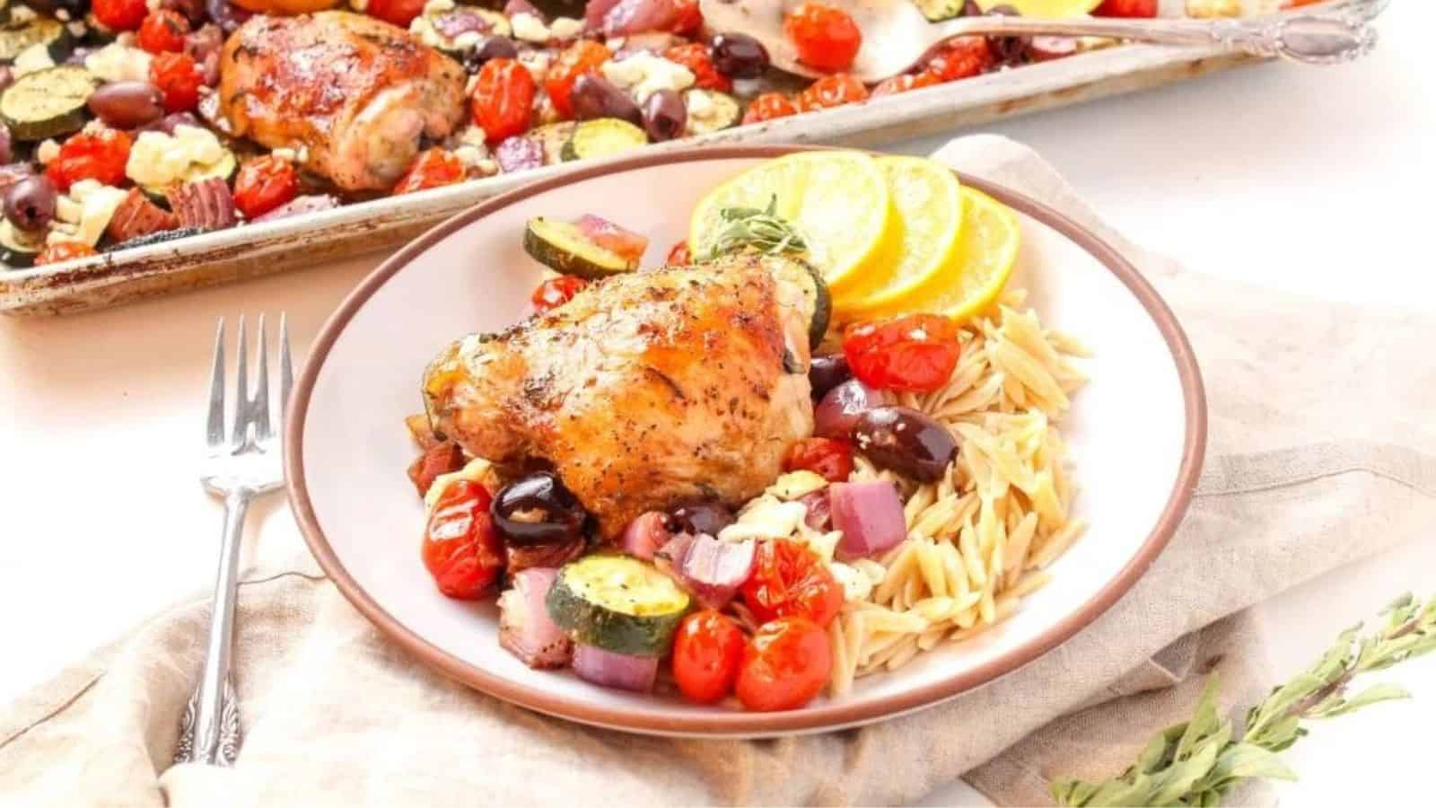 A shallow white bowl filled with a roasted chicken thigh, roasted zucchini, cherry tomatoes, red onion, kalamata olives, feta cheese, and orzo.