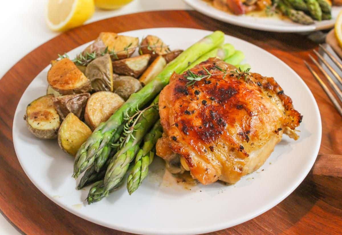 A cooked chicken thigh next to roasted asparagus and baby potatoes on a white plate.