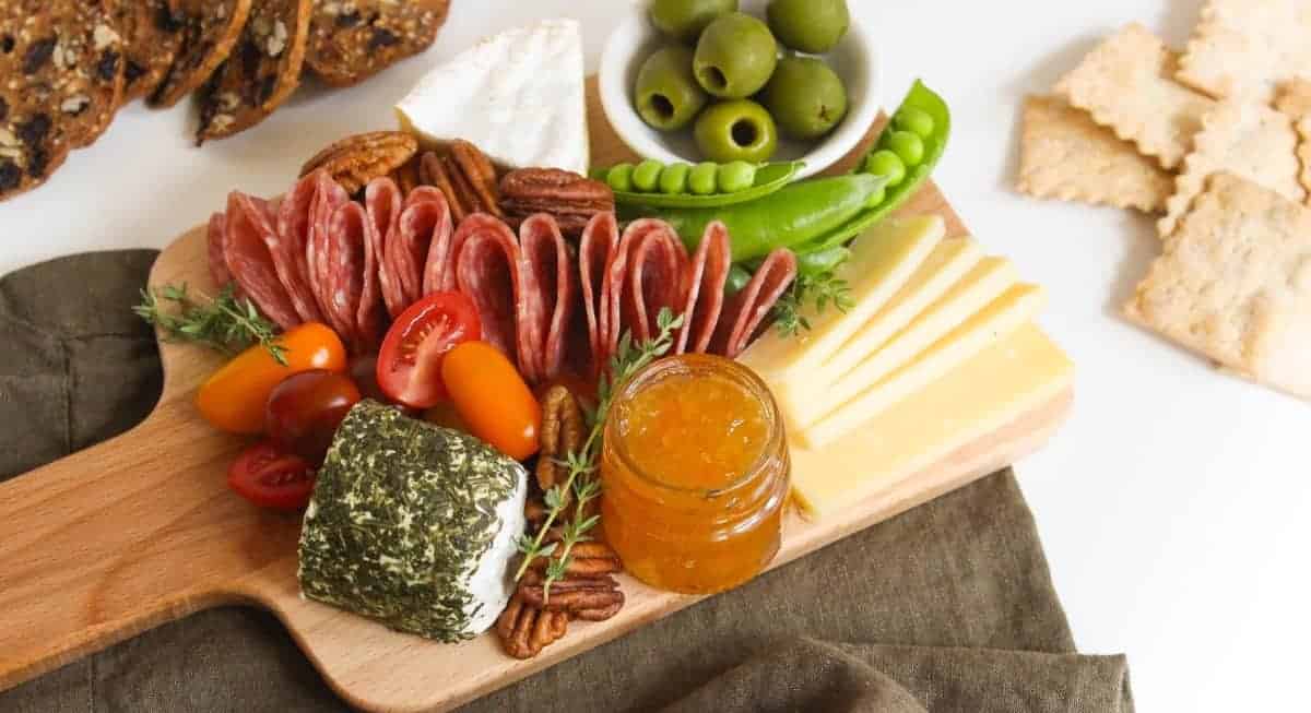 A small wooden board topped with cured meat, sliced cheese, dips, vegetables, nuts, and fruits.