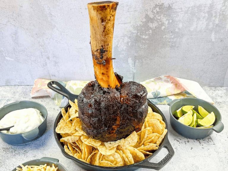 A large roasted meat shank on a bone, standing upright in a skillet surrounded by tortilla chips, with side dishes of lime wedges and sauce.