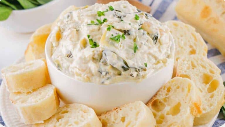 A bowl of spinach and artichoke dip garnished with chives, surrounded by sliced baguette on a blue patterned cloth.