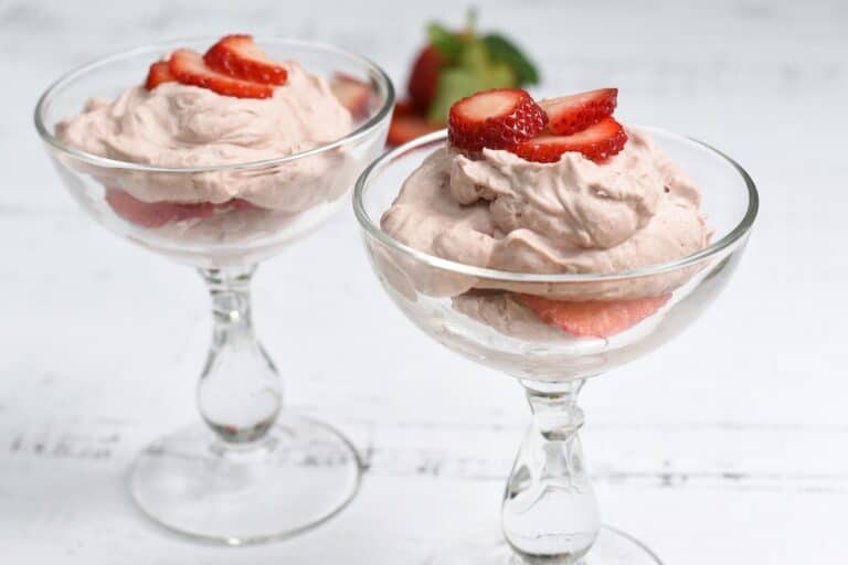 Two glasses of strawberry mousse topped with fresh strawberry slices, served on a white wooden surface.