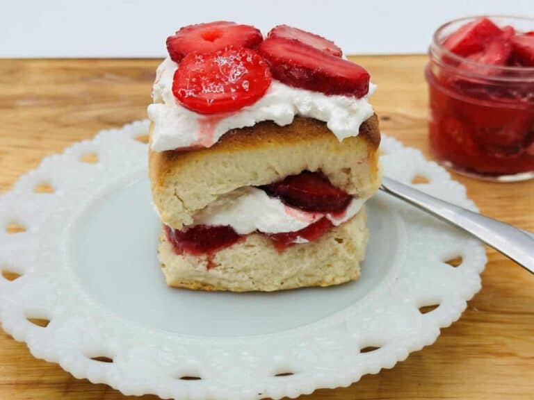 Strawberry shortcake with whipped cream served on a blue plate with a glass jar of strawberry jam in the background.