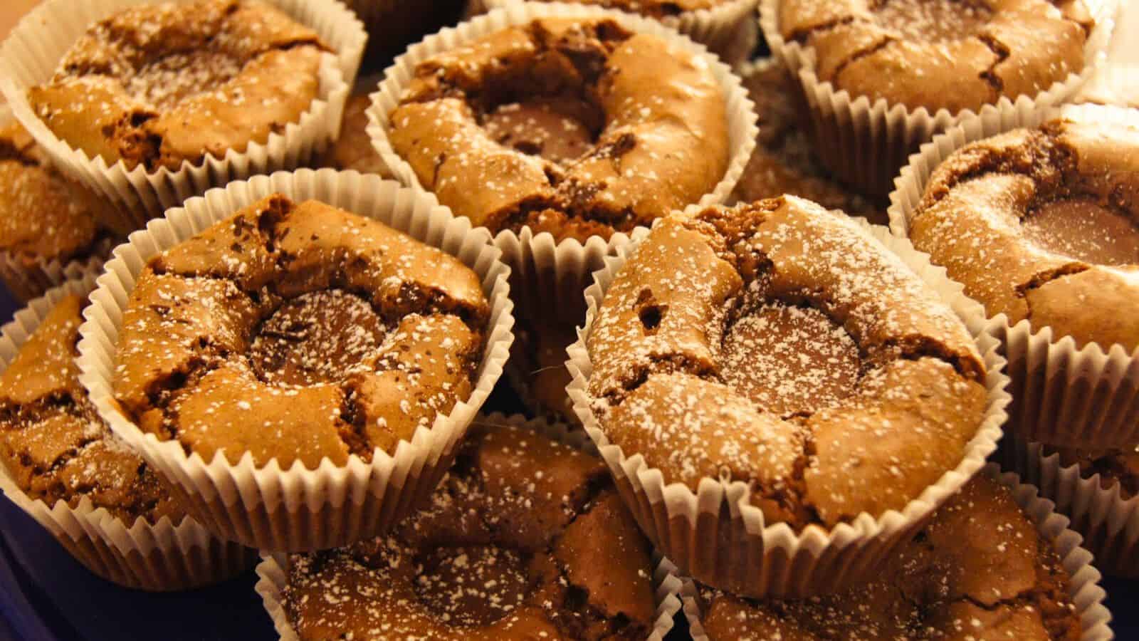 Image shows A close-up image of freshly baked brownies in muffin tins with a surprise in the middle and with a dusting of powdered sugar, placed in brown paper liners.
