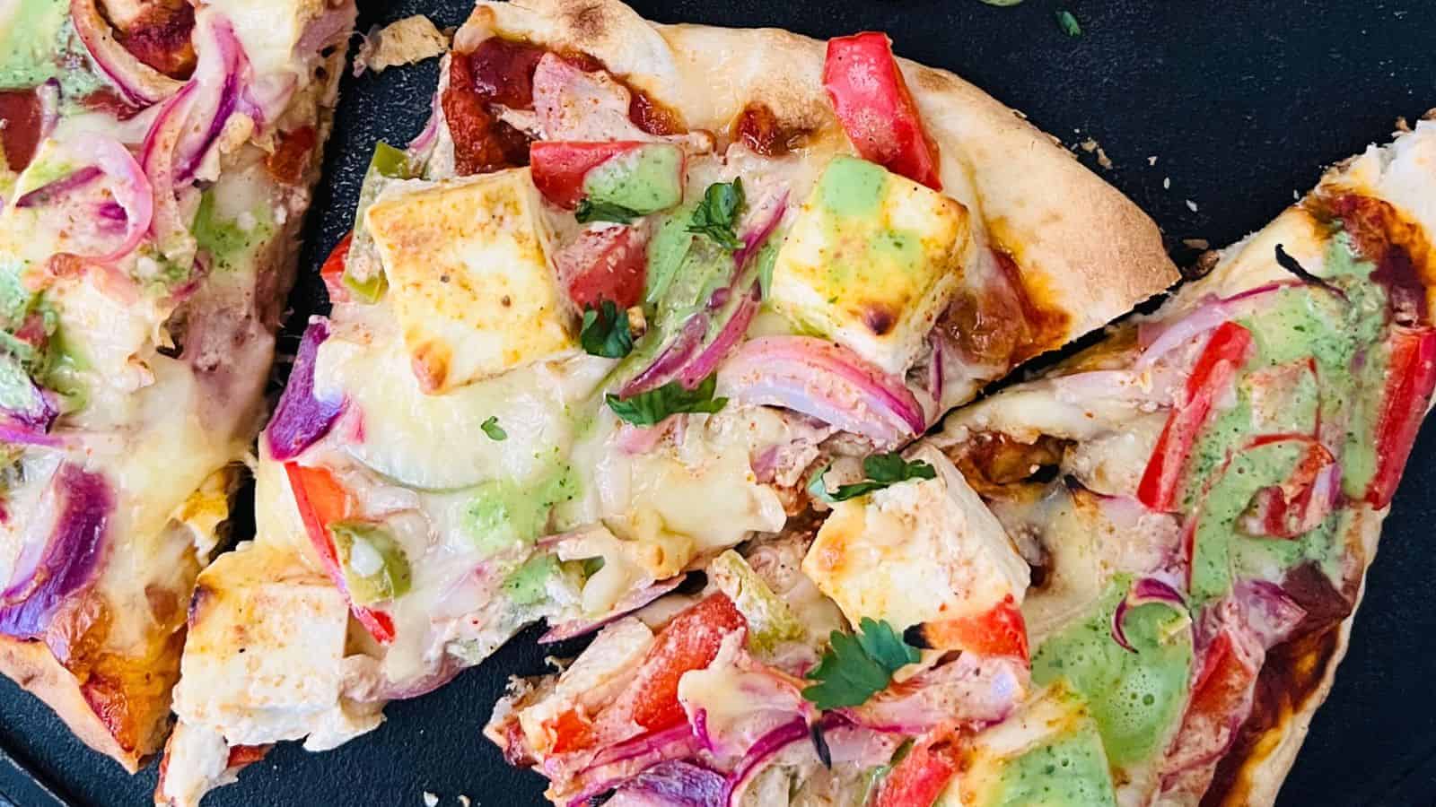 Slices of Tandori Paneer Naan Pizza with tofu, red onions, tomatoes, and drizzled with green sauce on a black surface.