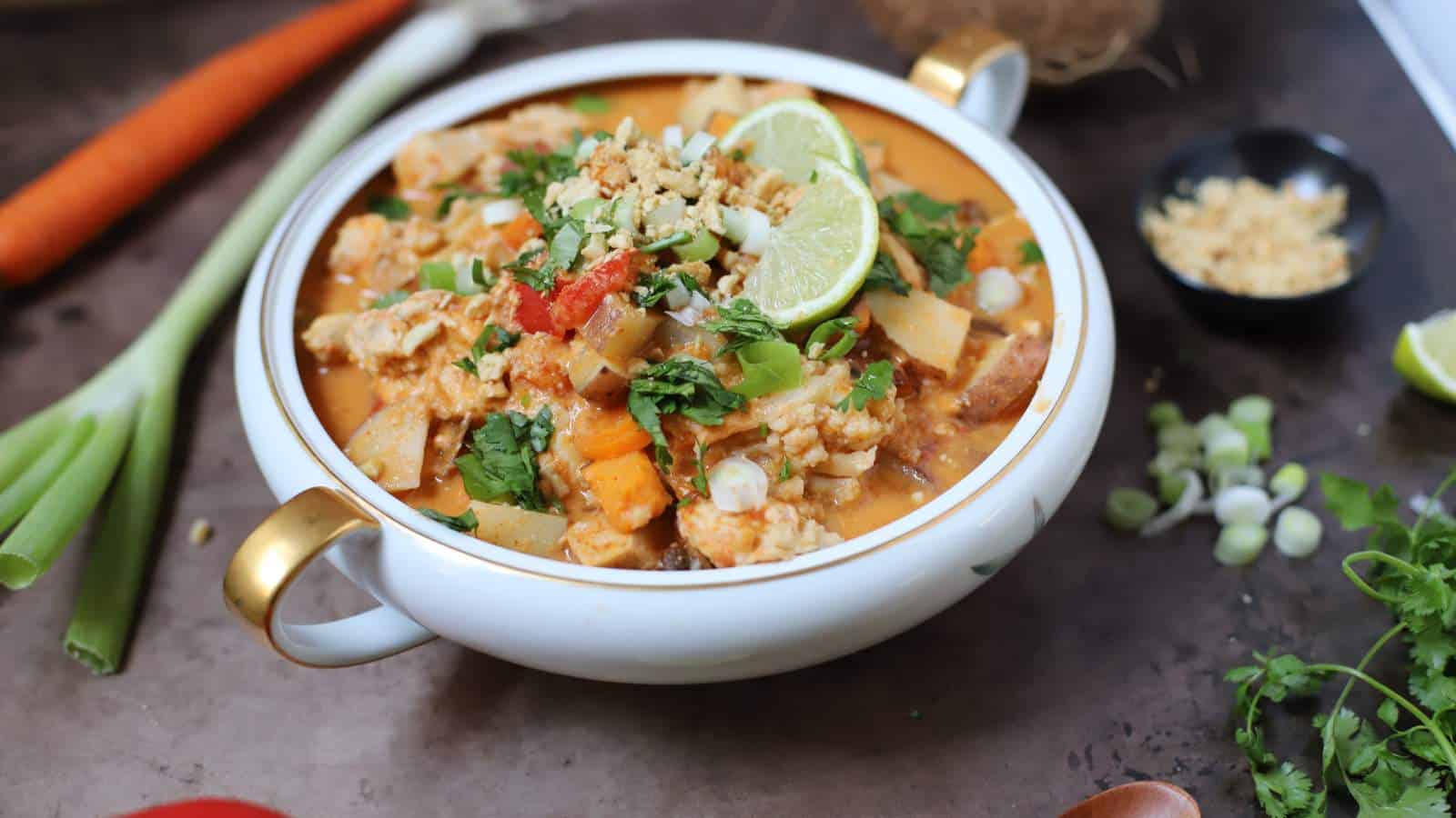 Thai chicken curry in a white bamboo bowl with a coconut and other fresh ingredients around it on a slate table.