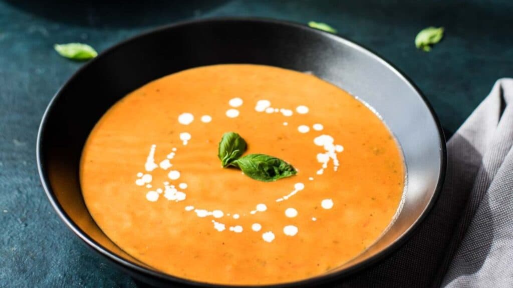 A bowl of creamy tomato soup garnished with fresh basil leaves and a swirl of cream.