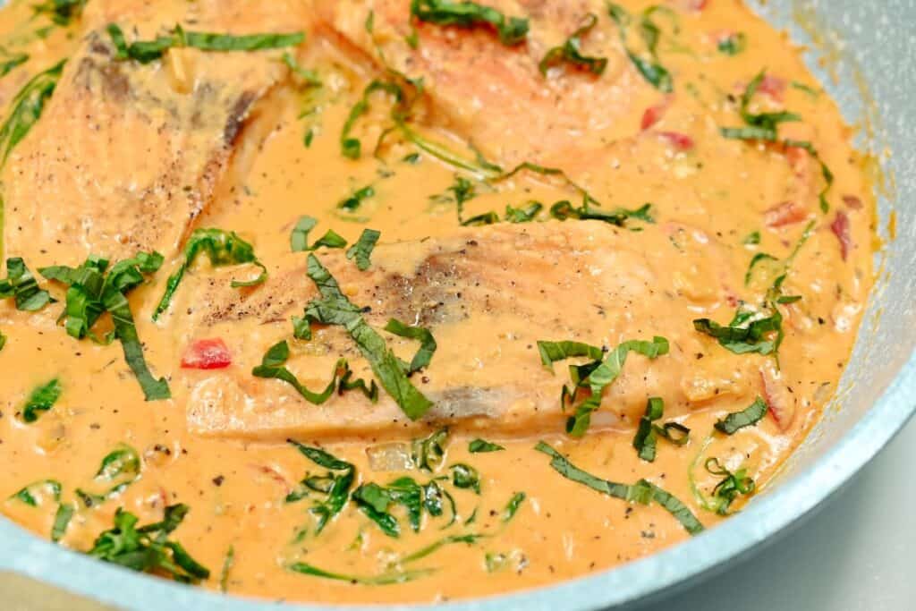 Salmon fillets cooking in a creamy sauce with herbs and spices in a skillet.