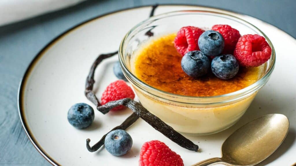 A serving of crème brûlée garnished with fresh raspberries and blueberries, accompanied by vanilla pods, on a ceramic plate with a spoon.