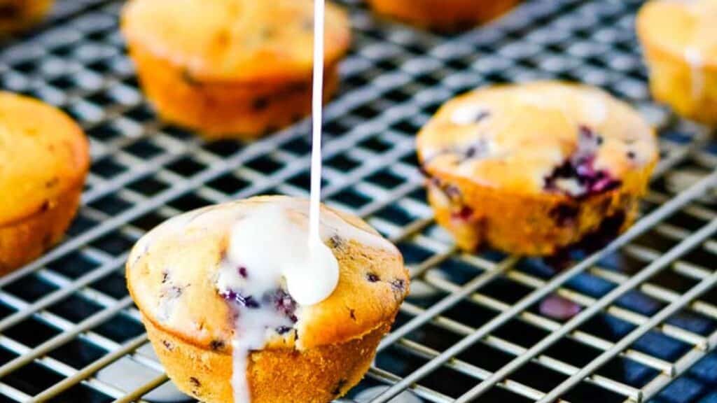 Freshly baked blueberry muffins on a cooling rack with icing being drizzled on top.