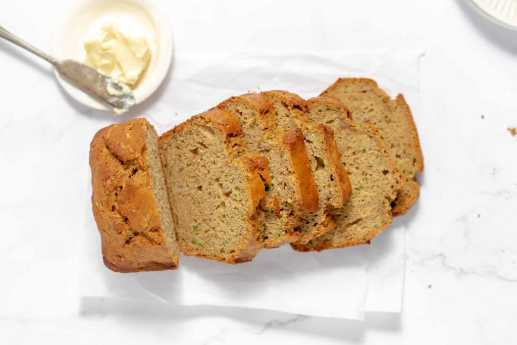 Sliced loaf of banana bread on white paper with a butter knife and dish in the background on a marble surface.