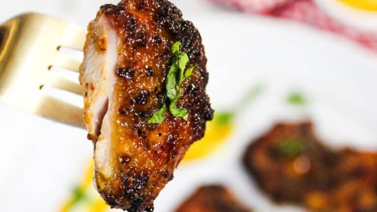 A piece of air fried chicken thigh on a fork, with a caramelized exterior and garnished with a small leaf, in focus against a blurred background of more chicken pieces.