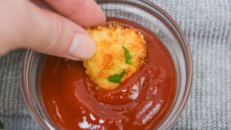 A hand dipping a breaded chicken nugget into a bowl of ketchup, garnished with a small basil leaf.