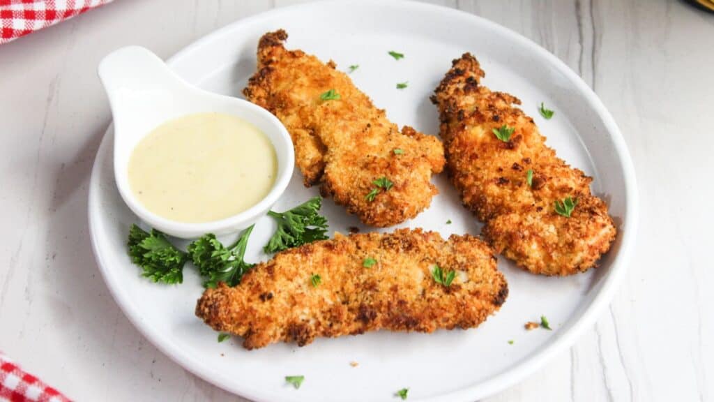 Crispy breaded chicken tenders on a white plate with a side of honey mustard sauce, garnished with parsley.