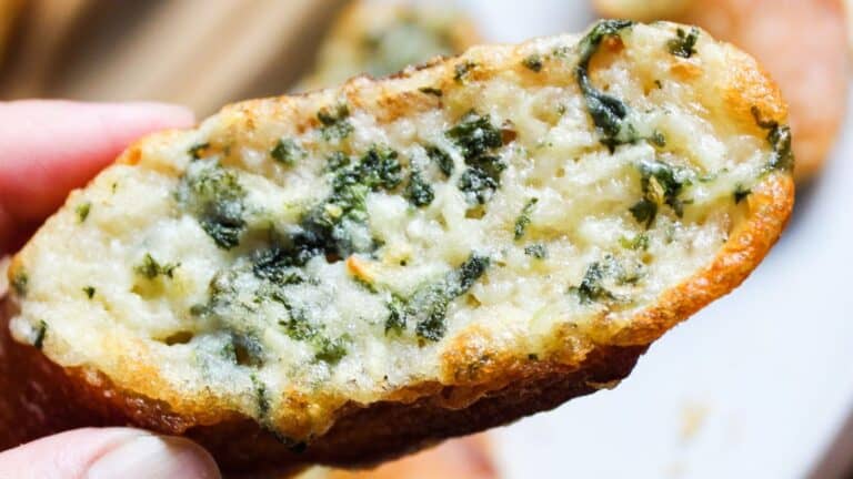 A close-up image of a hand holding a piece of garlic bread topped with melted cheese and herbs.