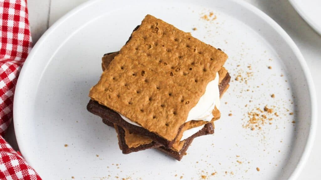 A s'more with melted marshmallow and chocolate between graham crackers on a white plate.