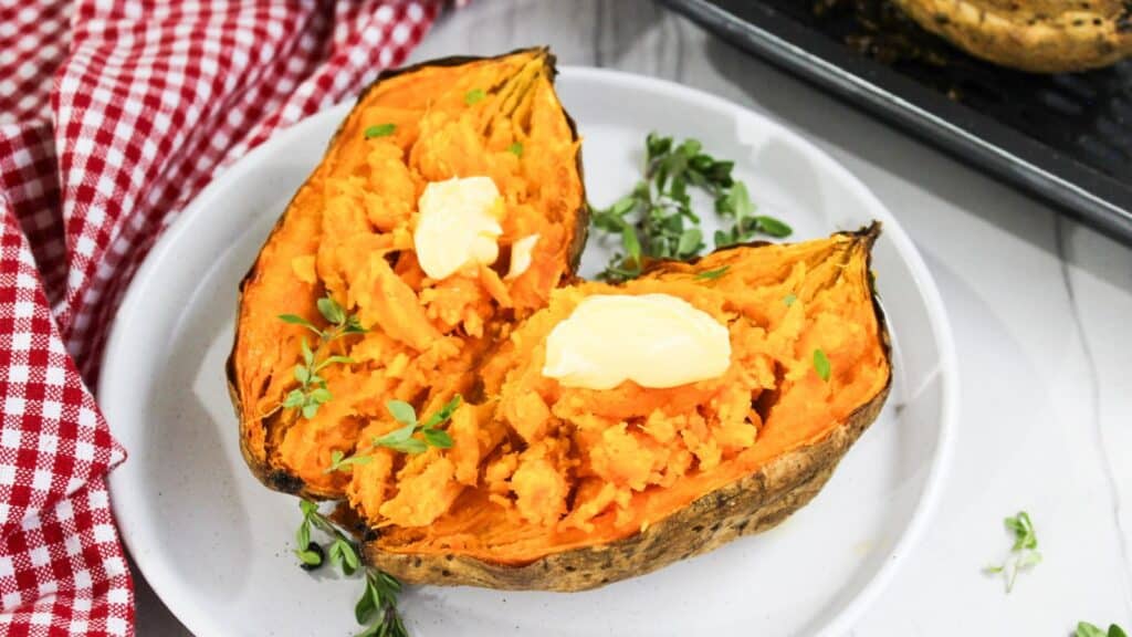 Two halves of a baked sweet potato with butter on a white plate, garnished with herbs, next to a checkered red napkin.
