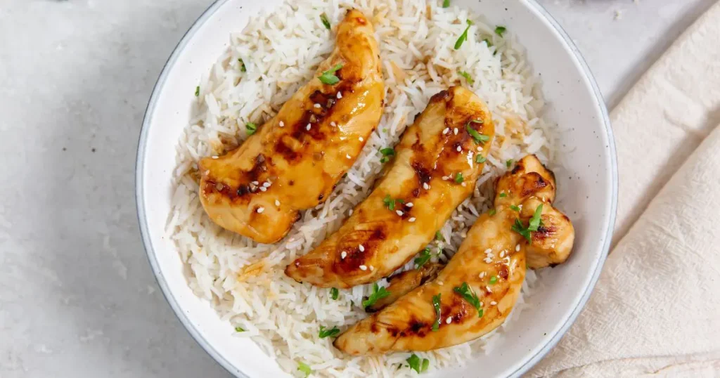 Three grilled chicken tenders glazed with a sweet sauce, served on a bed of white rice in a white bowl, garnished with chopped parsley.