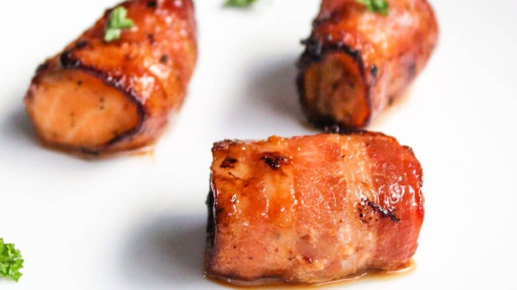 Three pieces of bacon-wrapped salmon bites garnished with parsley on a white plate.